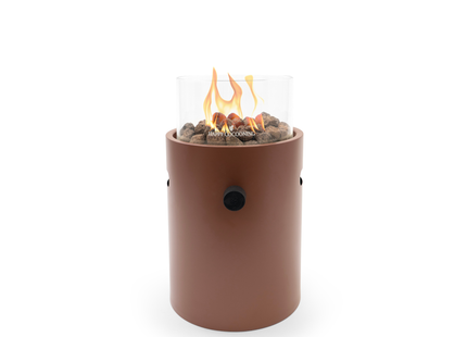 A modern outdoor fire pit with a coral red cylindrical body, clear glass wind guard, and visible orange flames over brown decorative stones. The brand name is printed on the front of the glass.
