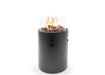 A modern cylindrical outdoor fire pit with a matte black body, clear glass wind guard, and visible orange flames over brown decorative stones. The brand name 'Focalrite' is printed on the front of the glass.