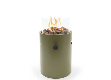 A modern outdoor fire pit with an olive green cylindrical body, clear glass wind guard, with flame on brown decorative stones. The brand name is printed on the front of the glass.