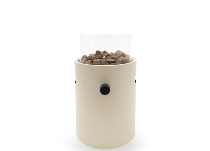 A modern cylindrical outdoor fire pit with a matte cream-colored body, a clear glass wind guard, and dark brown wood pellets, shown on a solid white background.