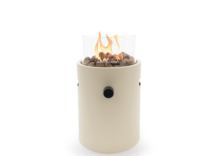 A modern cylindrical outdoor fire pit with a cream-colored body, a clear glass wind guard, dark stones, and a visible orange and yellow flame, set against a plain white background.